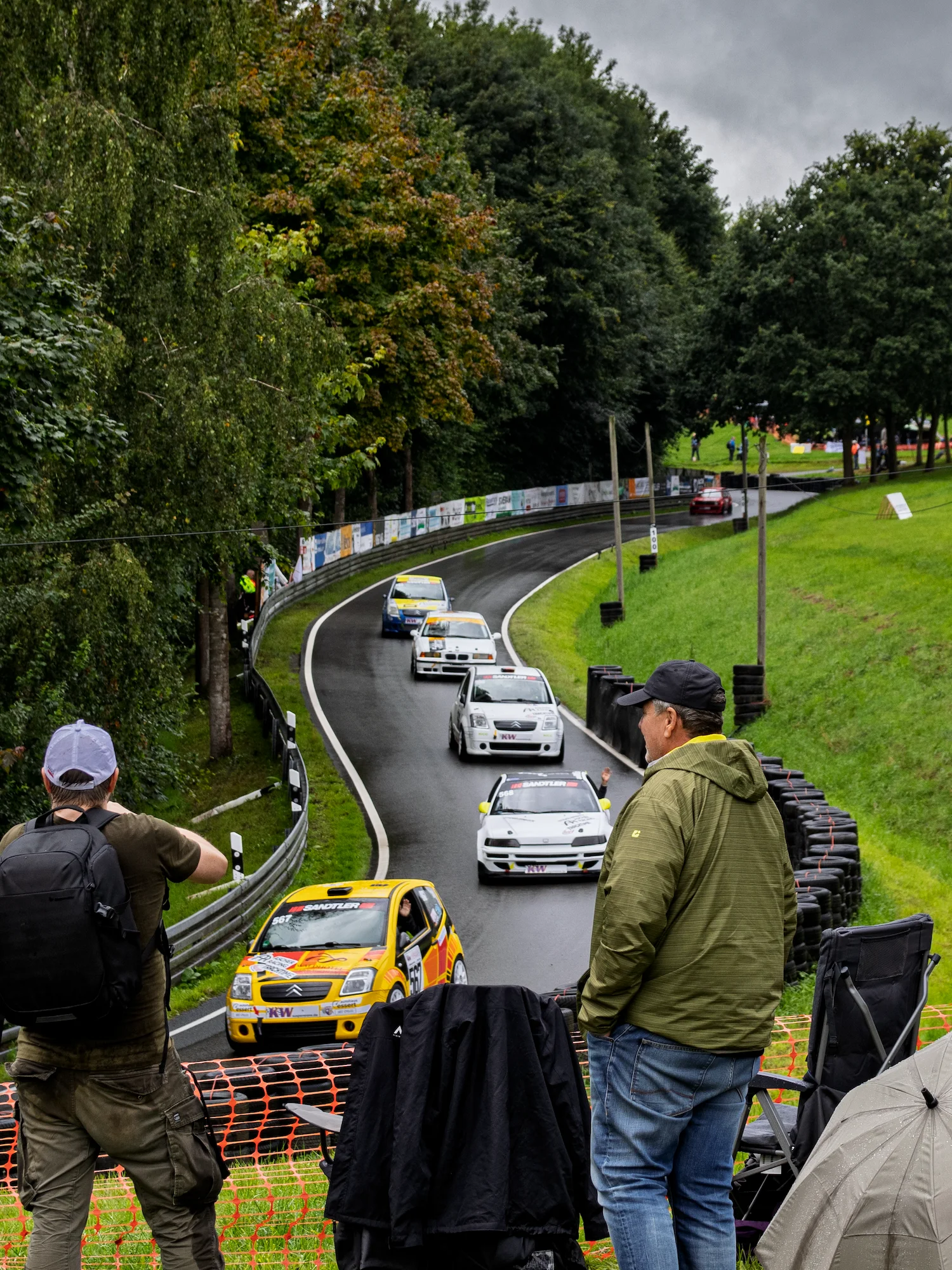 Fahrzeuge und Besucher des 57. Bergrennen auf einer Wiese an der Stecke und Fahrzeuge auf der Rennstrecke