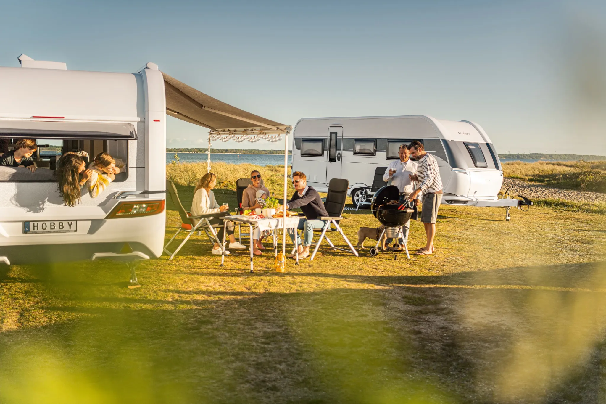 Familien grillen zwischen zwei Hobby Wohnwagen auf einer Wiese am See.