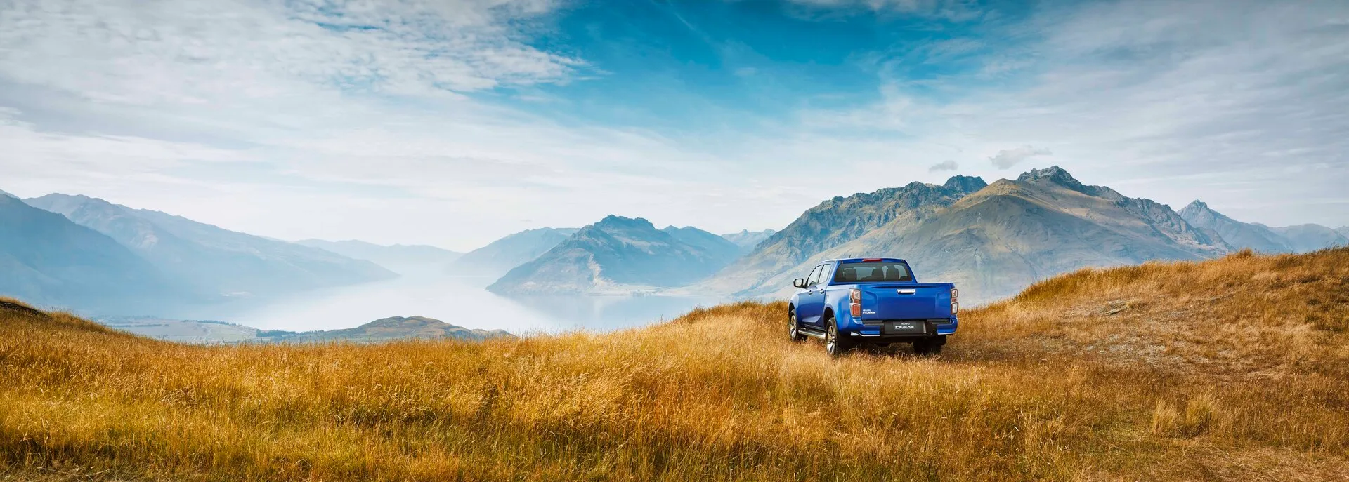 „Ein blauer Isuzu-Pick-up steht auf einer goldenen Wiese vor einer Bergkulisse unter blauem Himmel.“