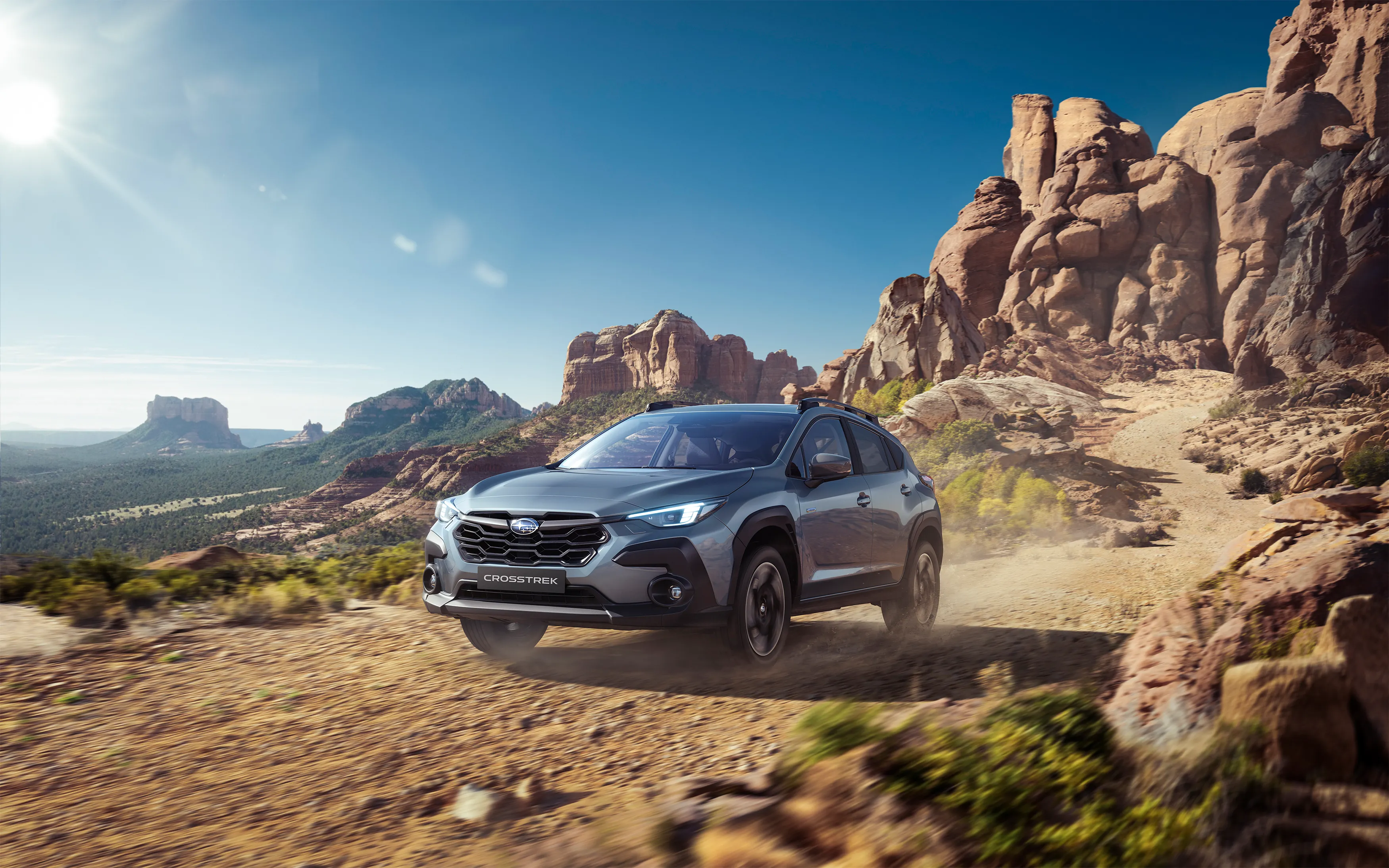 Blau- grauer Subaru Crosstrek fährt auf staubiger Piste durch felsige Wüstenlandschaft bei Sonnenschein.