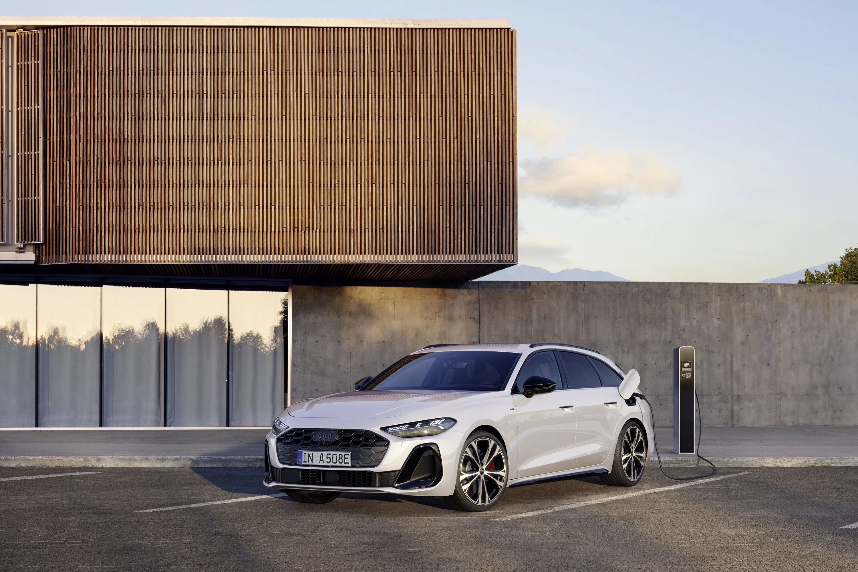 Weißer Audi A5 Avant Plug-in-Hybrid an einer Ladesäule vor moderner Architektur, mit sportlichem Exterieur, LED-Scheinwerfern und markanten Felgen – nachhaltiges Laden im Alltag.