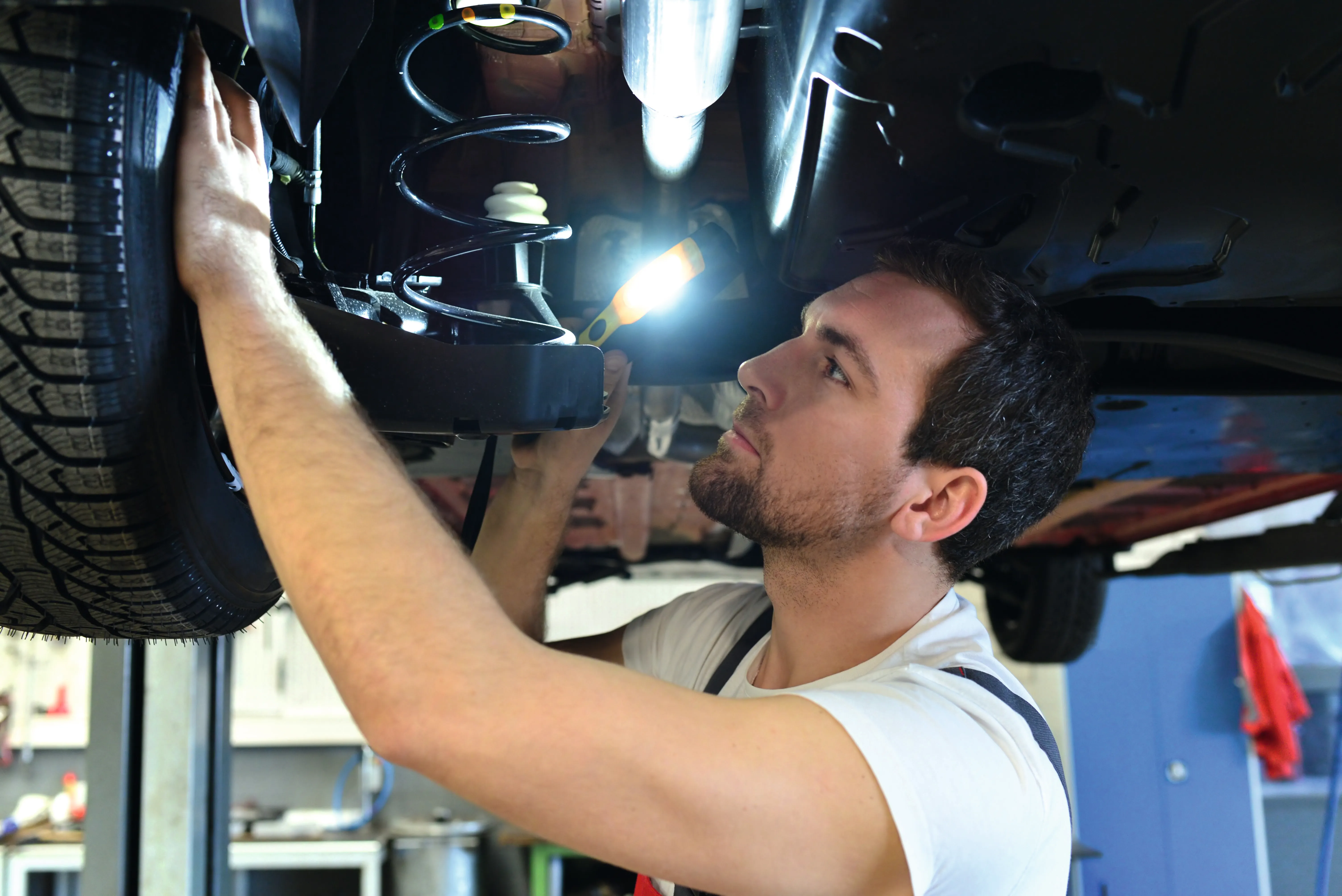 Ein Mechaniker führt unter einem aufgebockten Auto mit einer Lampe eine Sichtprüfung der Bremsanlage durch.