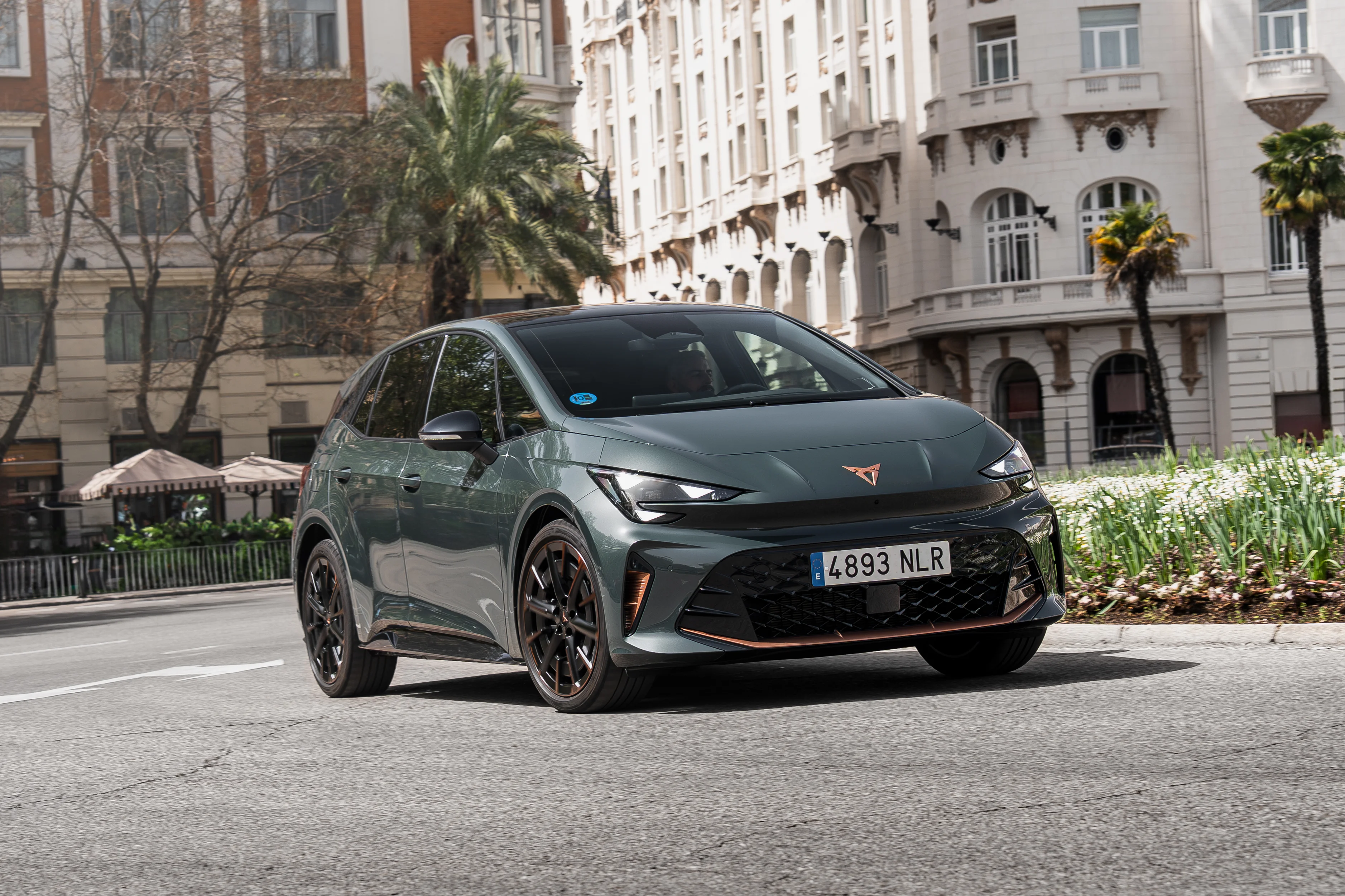 Grüner CUPRA Born VZ in der Farbe Dark Forest fährt durch eine moderne Innenstadt mit sportlichem Elektrodesign, kupferfarbenen Details und schwarzen Leichtmetallfelgen.