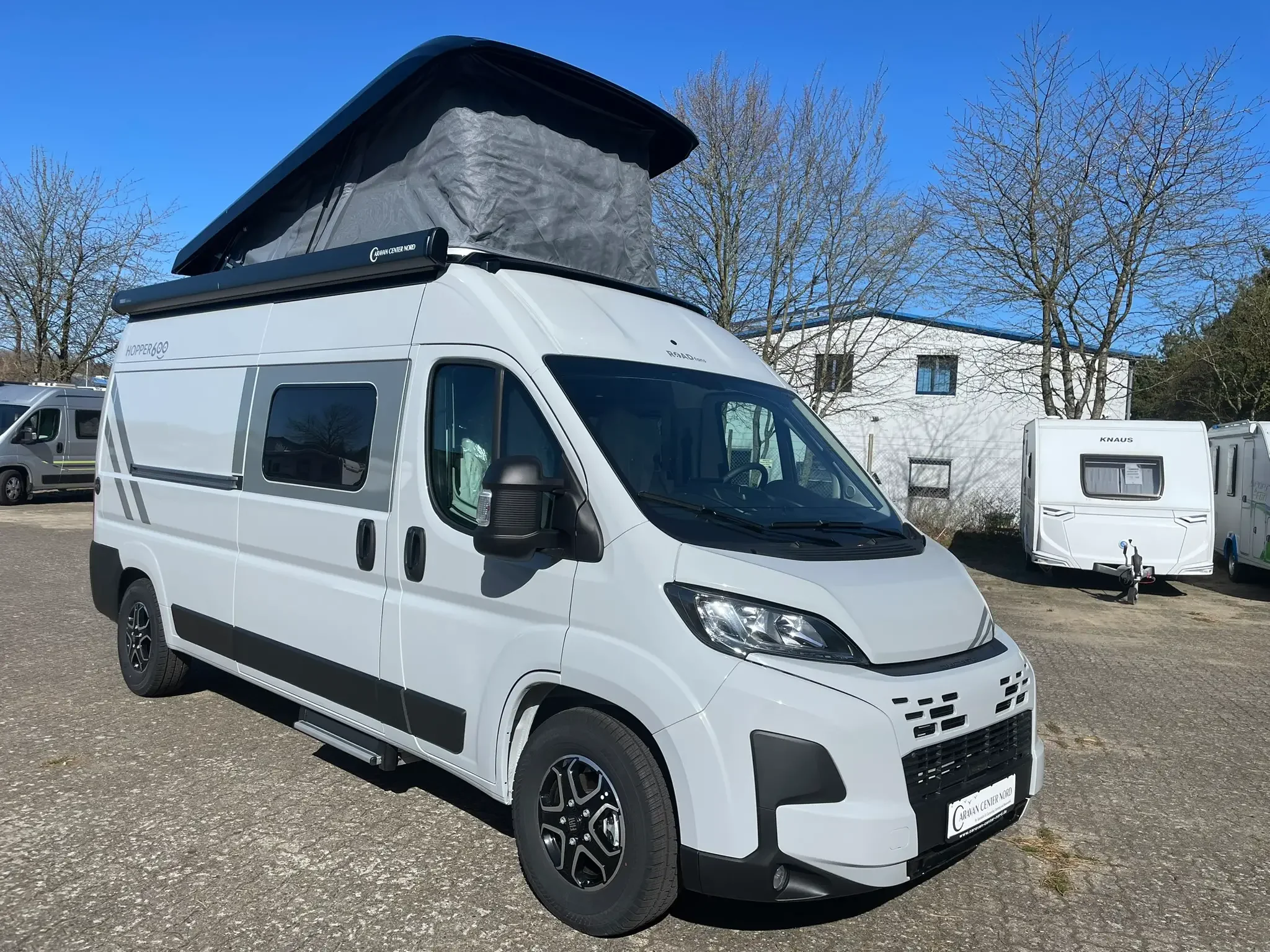 Weißer Camper Van der Marke RoadFans mit geöffnetem Aufstelldach auf Händlergelände – kompakter Kastenwagen für Camping und Reisen.