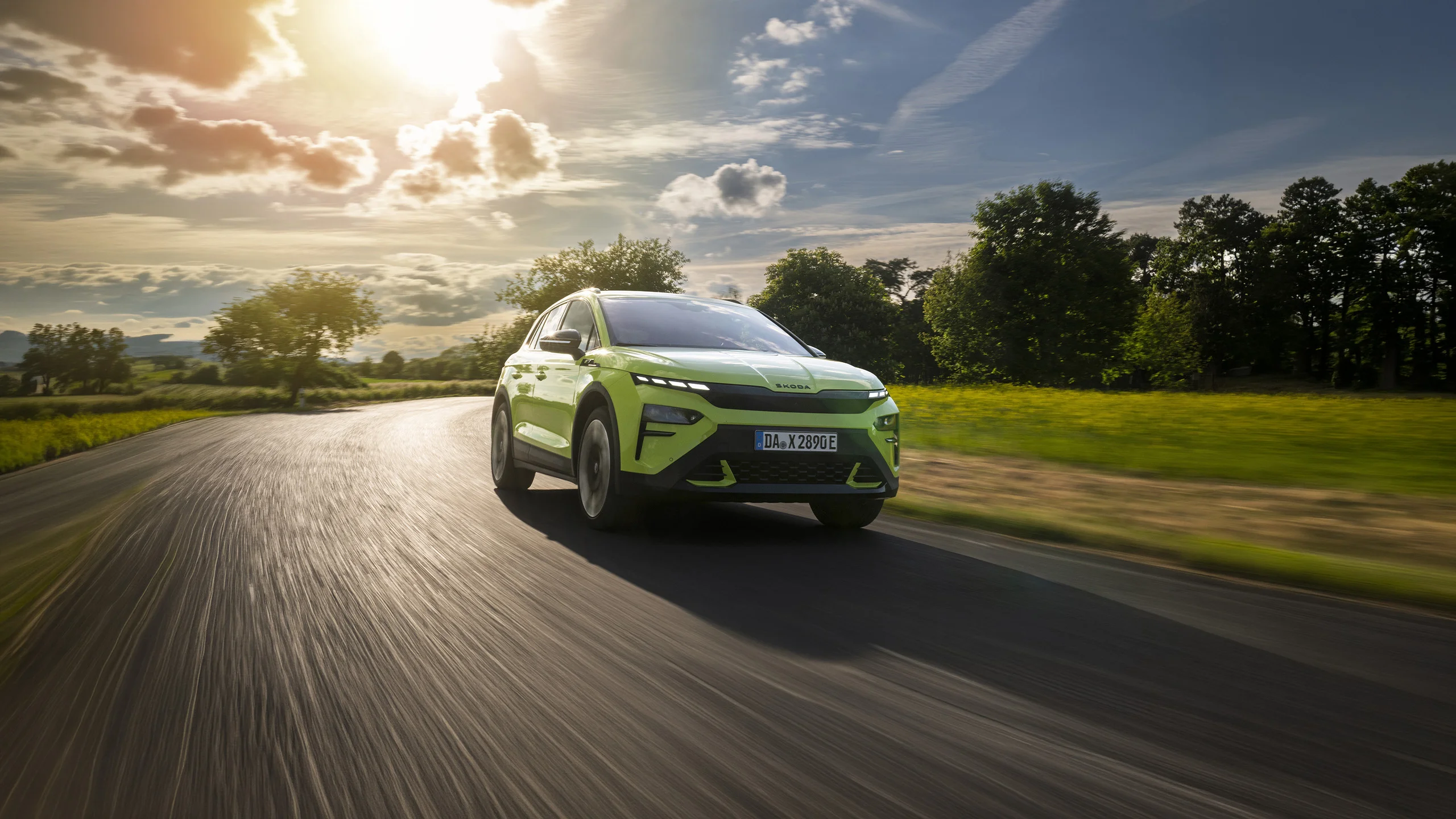 Skoda Elroq in Mamba-Grün während der Fahrt auf einer Straße zwischen Wiesen und Wäldern bei strahlendem Sonnenschein