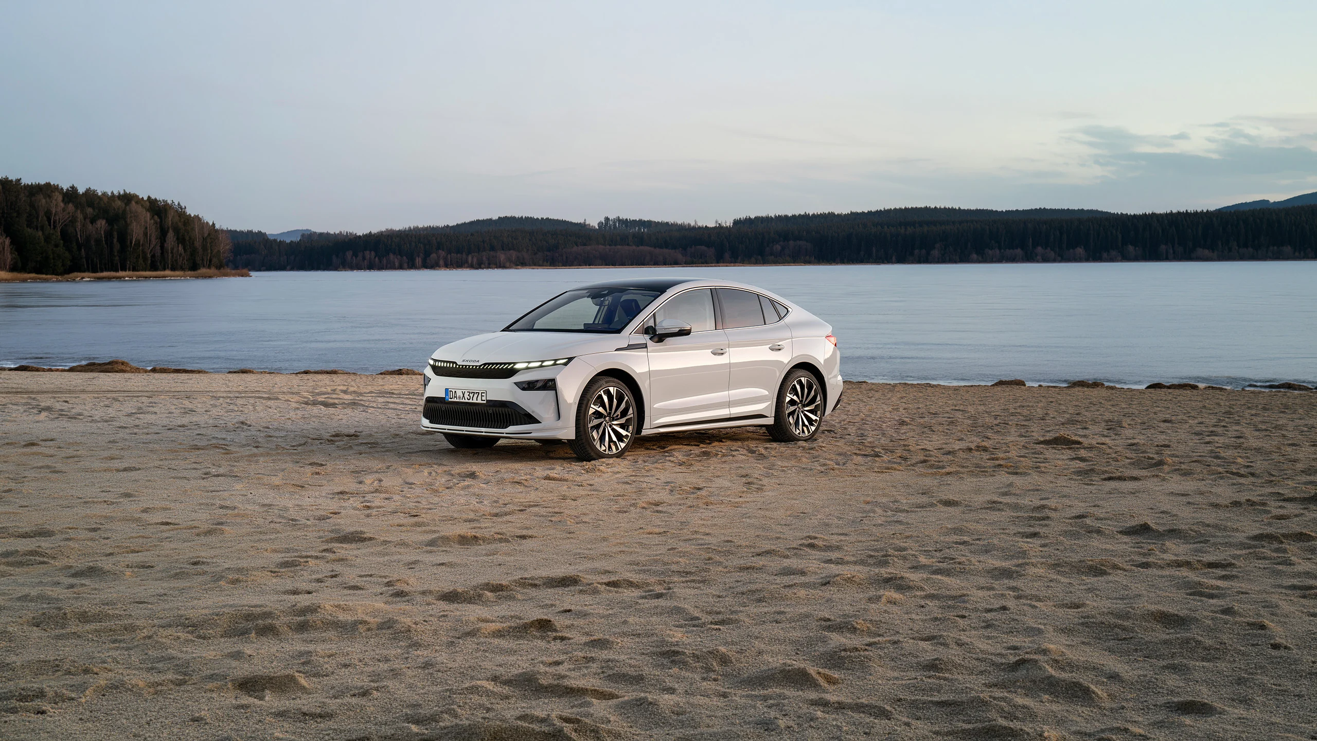 Der Skoda Enyaq Coupé in weiß an einem Sandstrand eines ruhigen Flusses mit Wald im Hintergrund