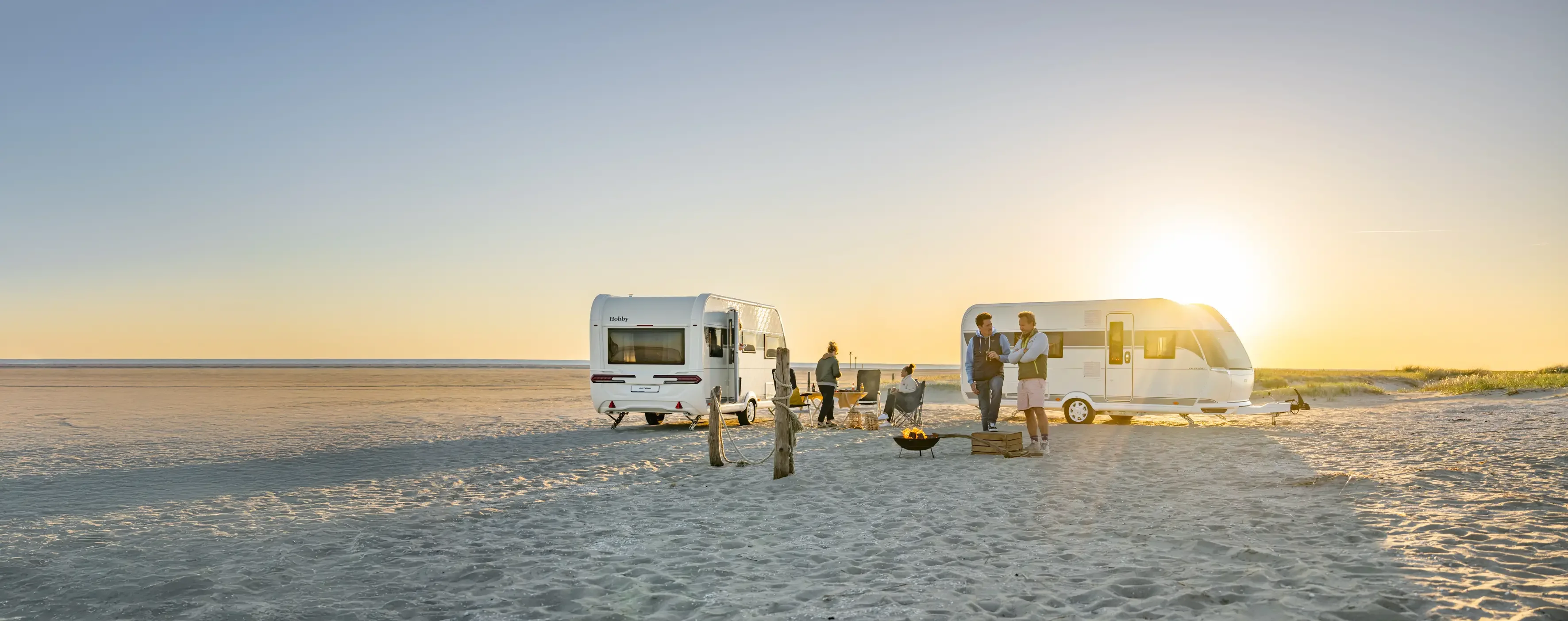 Wohnwagen am Strand bei Sonnenuntergang mit Menschen am Lagerfeuer – Campingurlaub an der Nordsee.